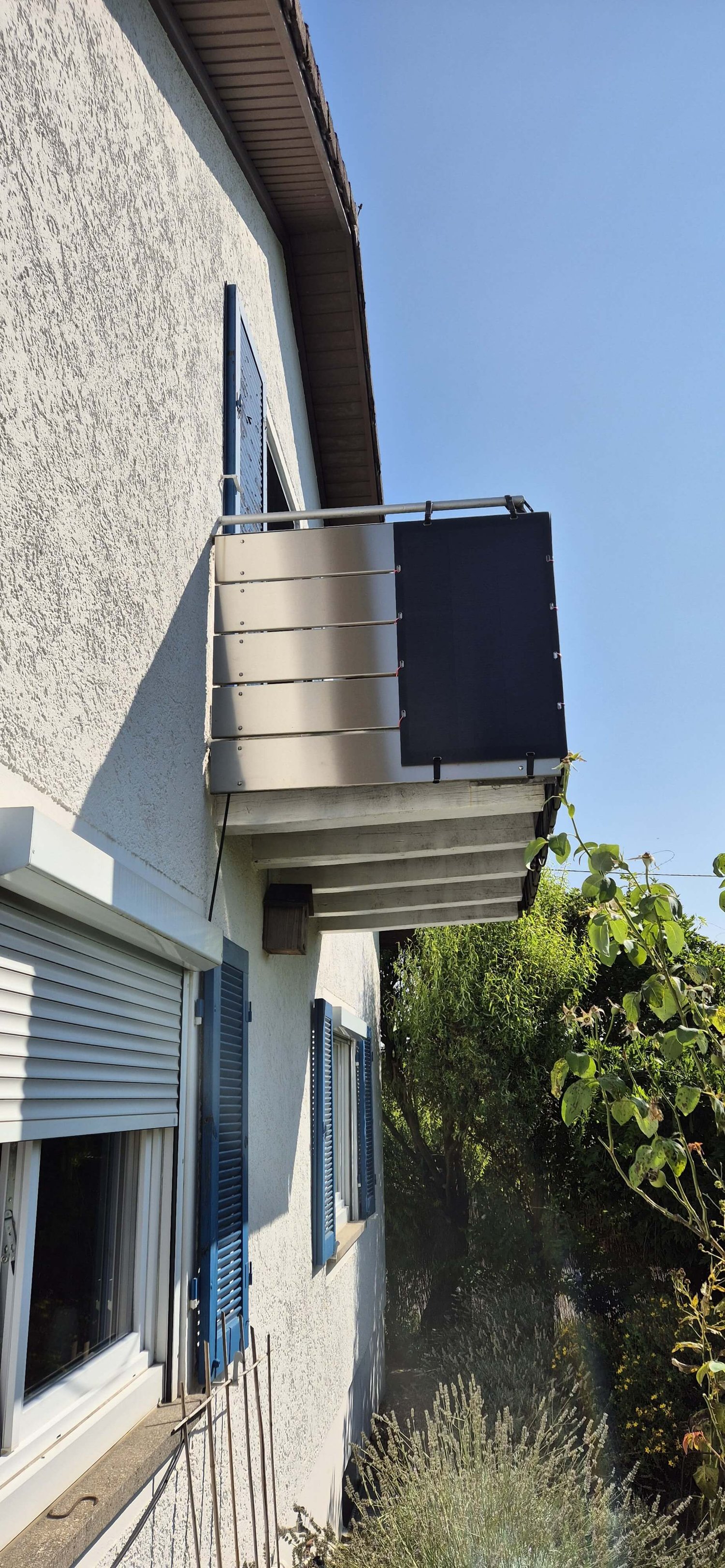 An der Seite eines Hauses mit weißen Wänden und blauen Fensterläden erstreckt sich ein Metallbalkon mit Sichtschutz und Solarpanel.
