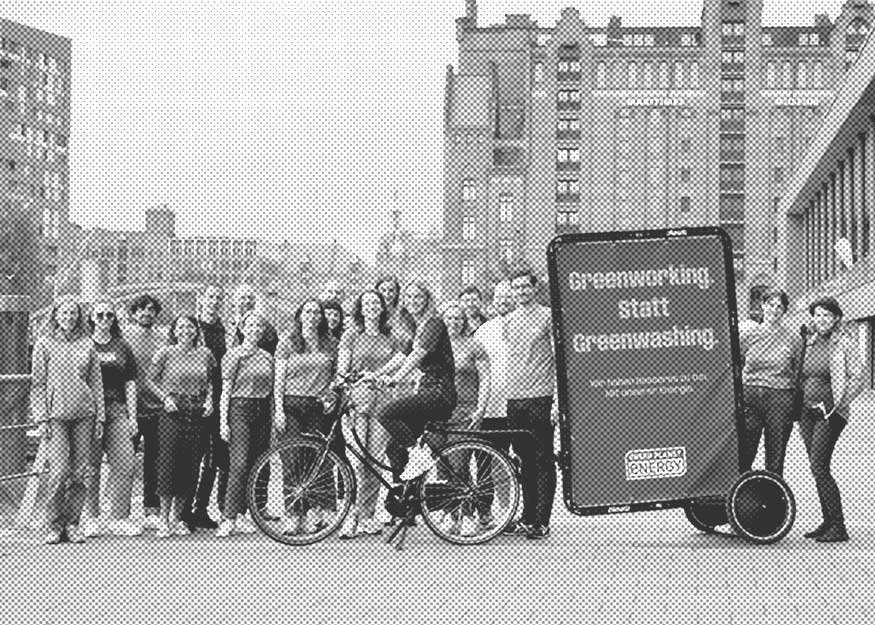 Eine Gruppe von Menschen steht im Freien neben einem Fahrrad mit Lastenanhänger mit der Aufschrift „Greenworking statt Greenwashing“ vor einer städtischen Kulisse.