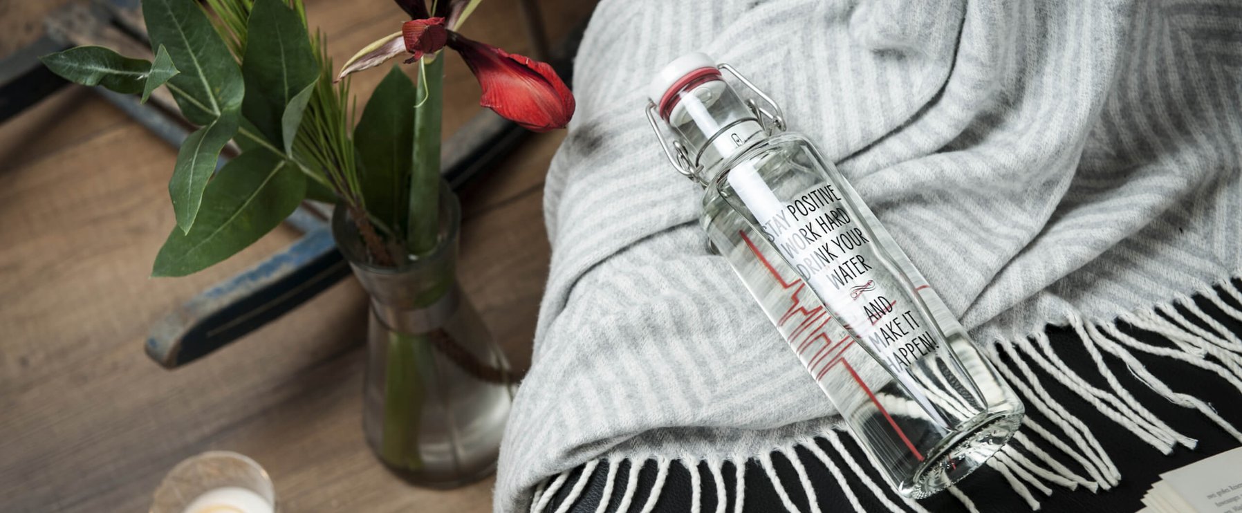 Eine Glasflasche mit rotem Muster auf einer grau gestreiften Decke, neben einer Vase mit grünen Blättern und einer roten Blume auf einer Holzoberfläche.