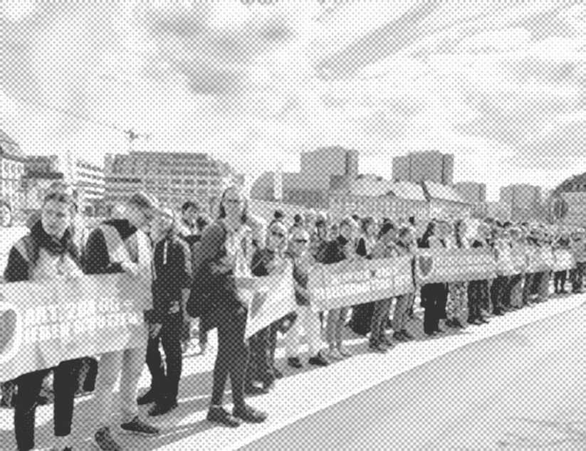 Eine Gruppe von Menschen marschiert mit einem langen Banner in der Hand zu einem Protestmarsch. Sie befinden sich im Freien, im Hintergrund sind Gebäude zu sehen.