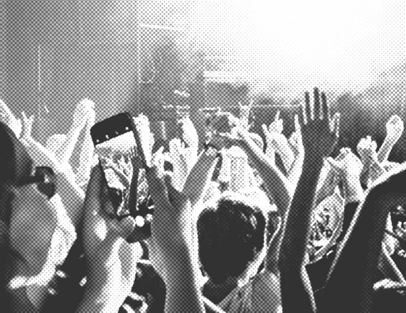 Bei einem Konzert hebt das Publikum die Hände. Ein Smartphone filmt die Darbietung. Die Szene ist in Schwarzweiß.