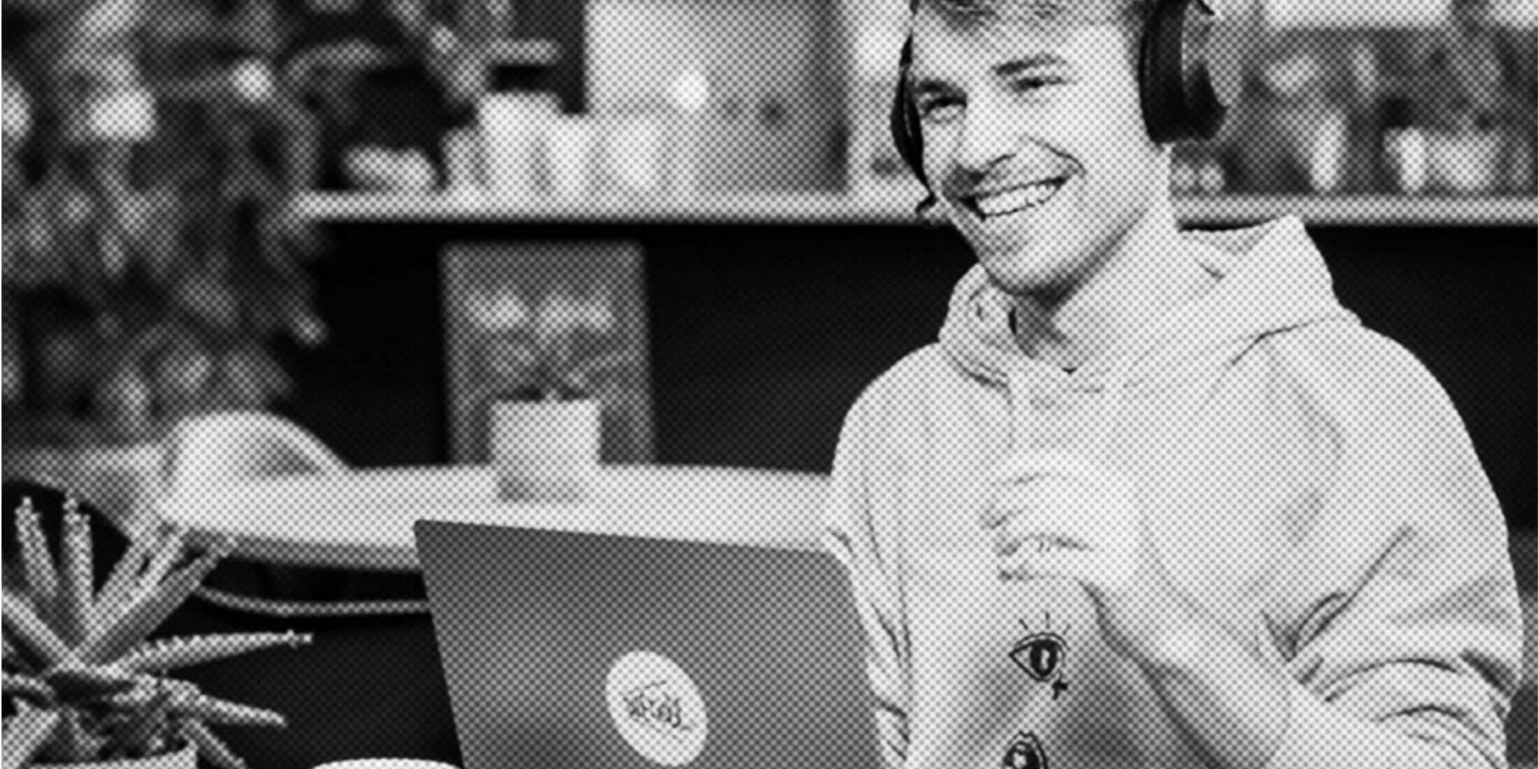 Eine Person im Kapuzenpullover lächelt, trägt Kopfhörer und benutzt einen Laptop in einem Raum mit Pflanzen und einem Tisch.